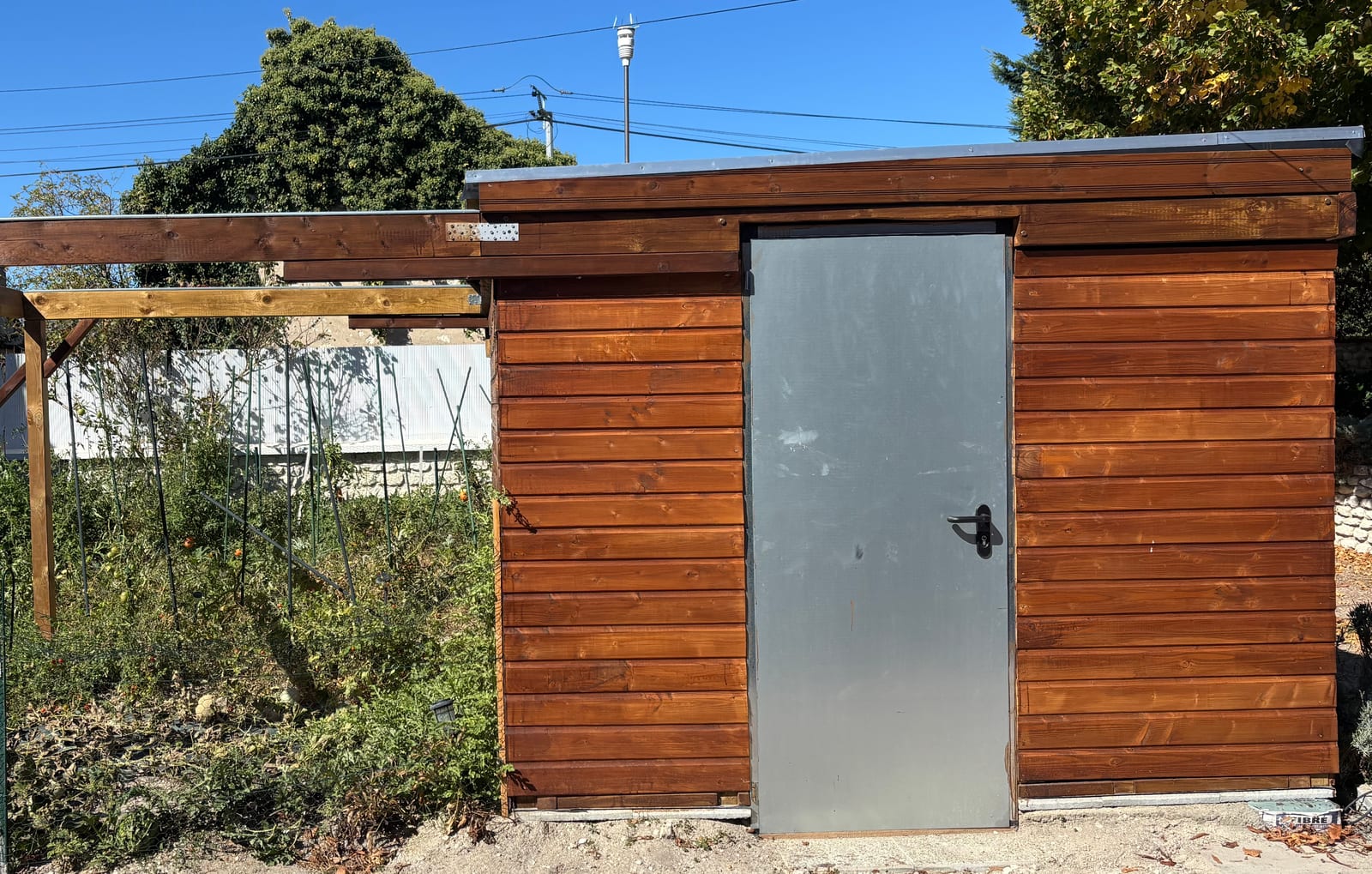 La façade est de l’abri, fermée, de jour : bardage bois, porte métallique, toit plat sous couvertine, et à gauche le portique sur poteaux où le toit vient se ranger, le mât de la station derrière.